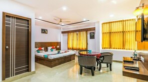 A hotel room with a bed, a desk, a chair, a window with curtains, and a ceiling fan. - Yuvraj Comforts (Mysore)