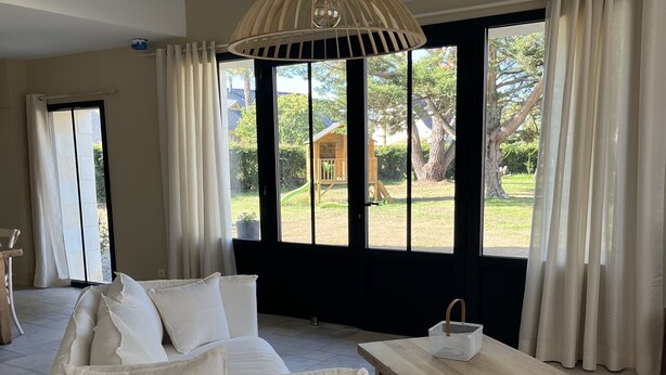 Un salón moderno con un sofá blanco, una mesa de centro de madera y vistas a un jardín a través de amplios ventanales.