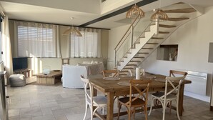 Amplia sala de estar con una mesa de comedor de madera, sillas blancas y una escalera que lleva a un nivel superior.