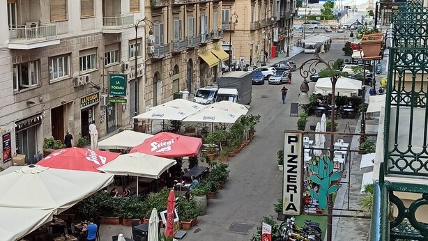Un carrer amb terrasses a l'aire lliure, plantes en testos i un cartell de pizzeria.
