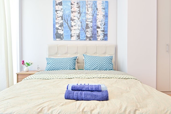 Una cama bien tendida con un edredón color beige y dos almohadas, un cuadro colgado en la pared con árboles de abedul, una mesita de noche con un florero y una mesita de noche con un reloj.