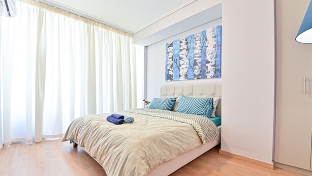Un dormitorio con cama, cabecera, un cuadro en la pared y ventana con cortinas.