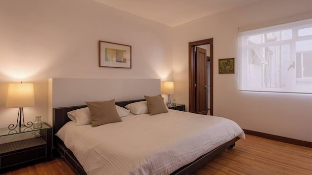 Une chambre avec un grand lit, deux lampes de chevet, un tableau encadré au mur et une fenêtre donnant sur l’extérieur.