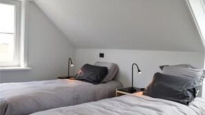 Une chambre avec deux lits, chacun étant doté d’une table de chevet et d’une lampe de bureau. Il y a une fenêtre sur le côté gauche, et la chambre a un plafond en pente.