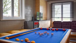 Une table de billard avec des billes orange et rouges, un canapé et un téléviseur dans une pièce aux murs en briques et aux grandes fenêtres.