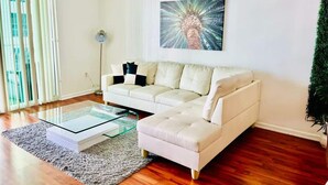 Een moderne woonkamer met een witte bank, een glazen salontafel en een groot schilderij aan de muur.