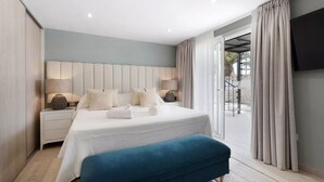 Quarto moderno com uma cama grande, duas mesas de cabeceira com abajures, um banquinho azul e uma TV na parede.