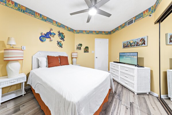 A bedroom with a bed, a ceiling fan, a TV, a dresser, and decorative wall art.