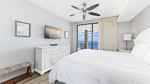 A hotel room with a large bed, a ceiling fan, a TV, a bench, a dresser, and a view of the ocean through a balcony door. - Summer House 1105A (Orange Beach)