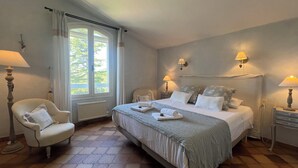 A bedroom with a large bed, two bedside lamps, a chair, a window with curtains, and a radiator. - Villa Maya : Large villa and amazing pool in Luberon (Mérindol)