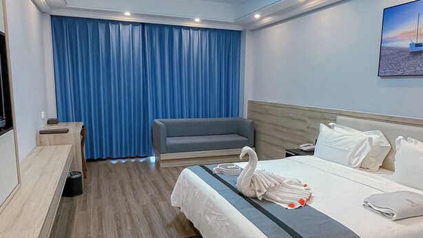 A modern hotel room with a wooden floor, a bed with white linens, a grey sofa, a desk with a chair, and a blue curtain. - G.C Royal Hotel (Accra)