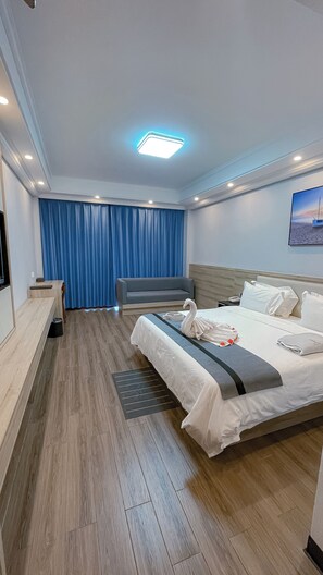 A modern hotel room with a wooden floor, a bed with white linens, a grey sofa, a desk with a chair, and a blue curtain. - G.C Royal Hotel (Accra)