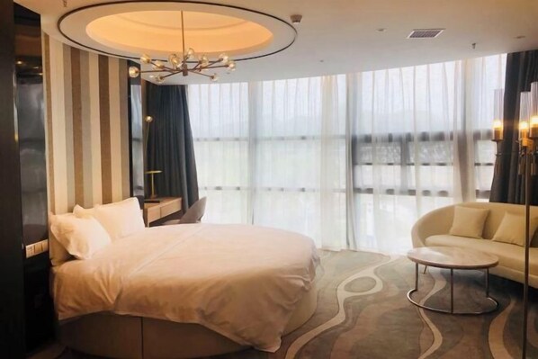 A hotel room with a large bed, a sofa, a round coffee table, and a view of the city through sheer curtains.