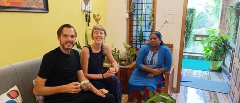Tres personas sentadas en un sofá en una habitación con una pared amarilla, una ventana con plantas y un tapiz decorativo.
