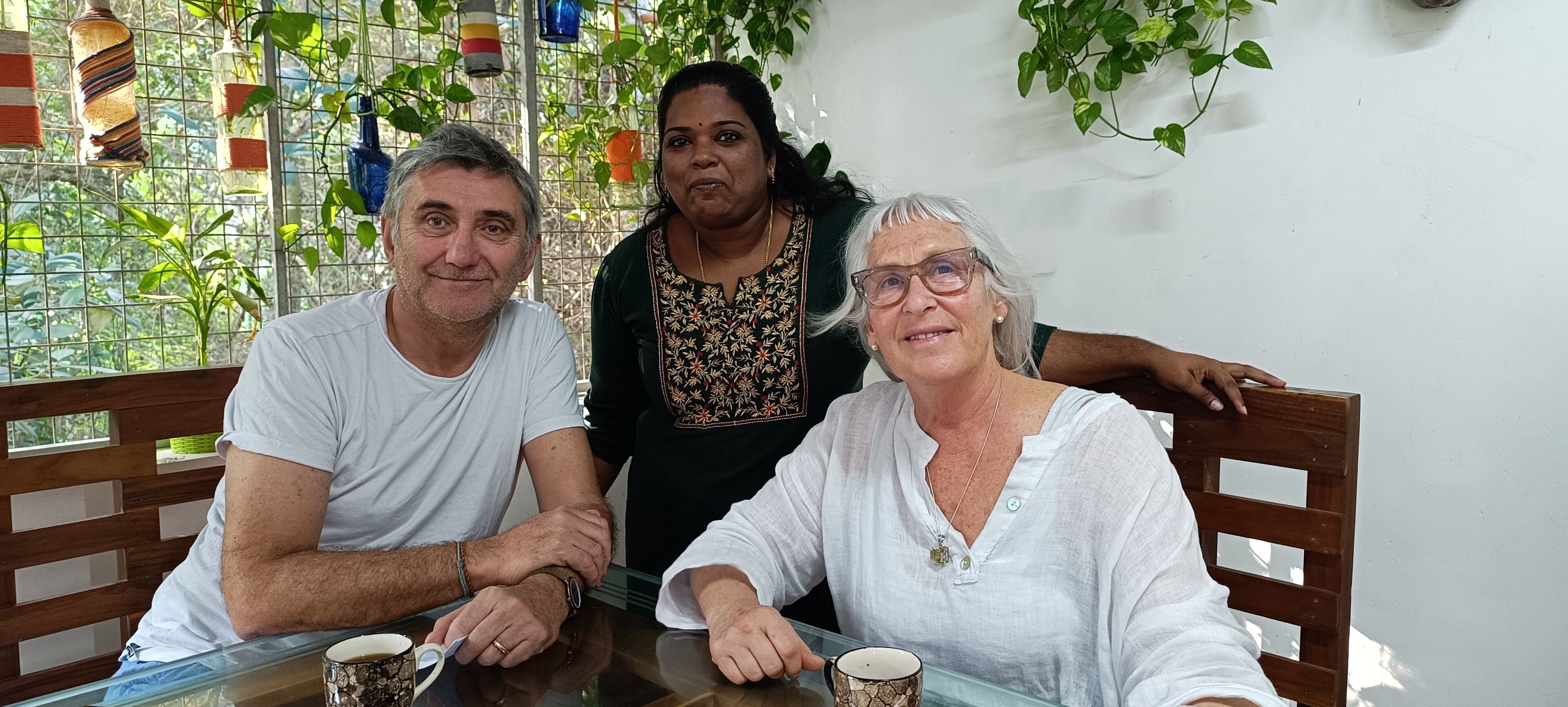 Three people seated at a table with a glass top, surrounded by greenery and a metal fence.