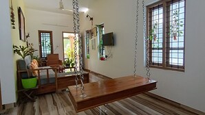 Een kamer met een houten schommel, een groene emmer, een houten bank, een aan de muur bevestigde televisie en een raam met planten.