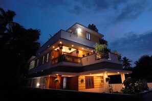 Une maison moderne à deux étages, dotée d’un balcon à l’étage supérieur, de nombreuses fenêtres et d’un éclairage extérieur.