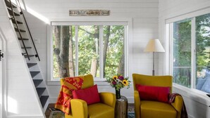 A room with a staircase, two yellow chairs with red cushions, a lamp, and a window with a view of trees. - Marion Lake Tree House (Perham)