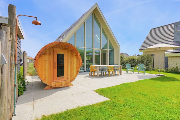 Une maison moderne dotée d’une grande baie vitrée, d’un sauna en bois et d’une terrasse avec des chaises.
