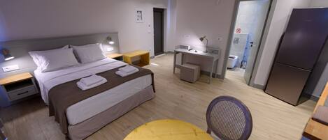 A modern hotel room with a bed, bedside tables, a desk, a chair, and a refrigerator.