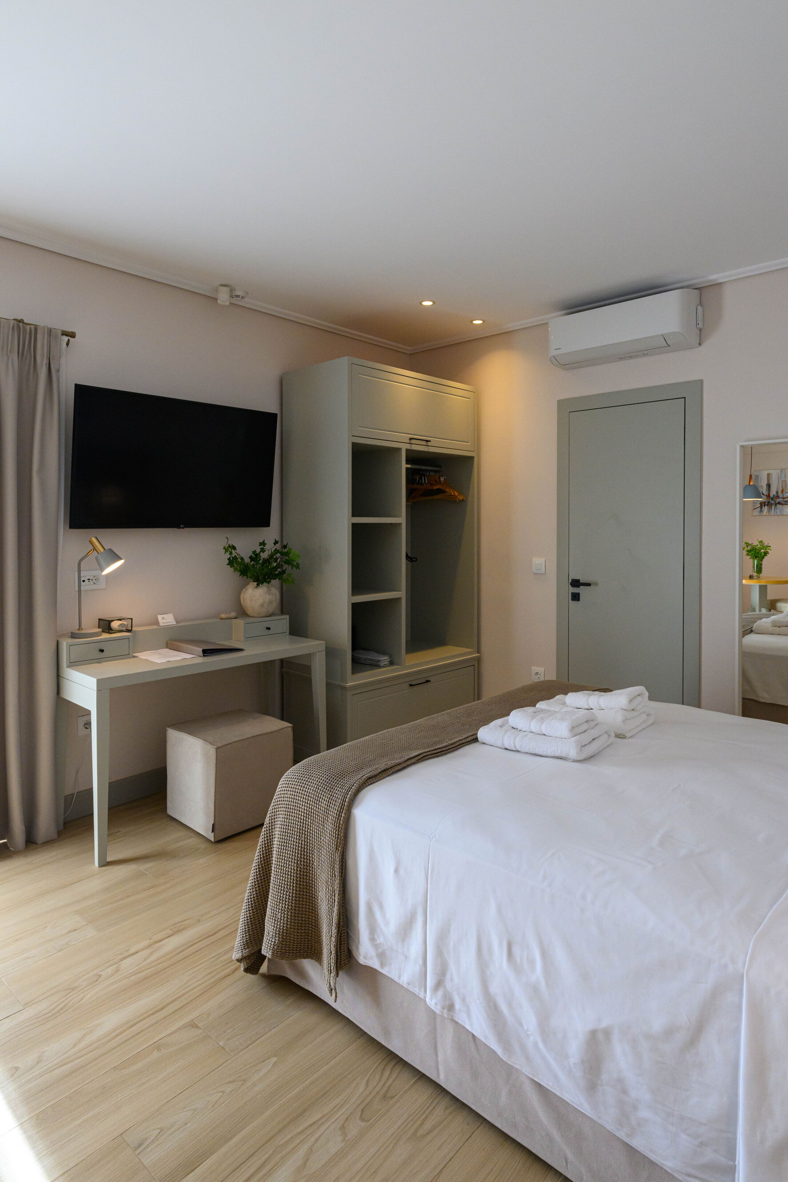 A modern hotel room with a bed, a desk with a lamp, a television, a wardrobe, and an air conditioning unit.