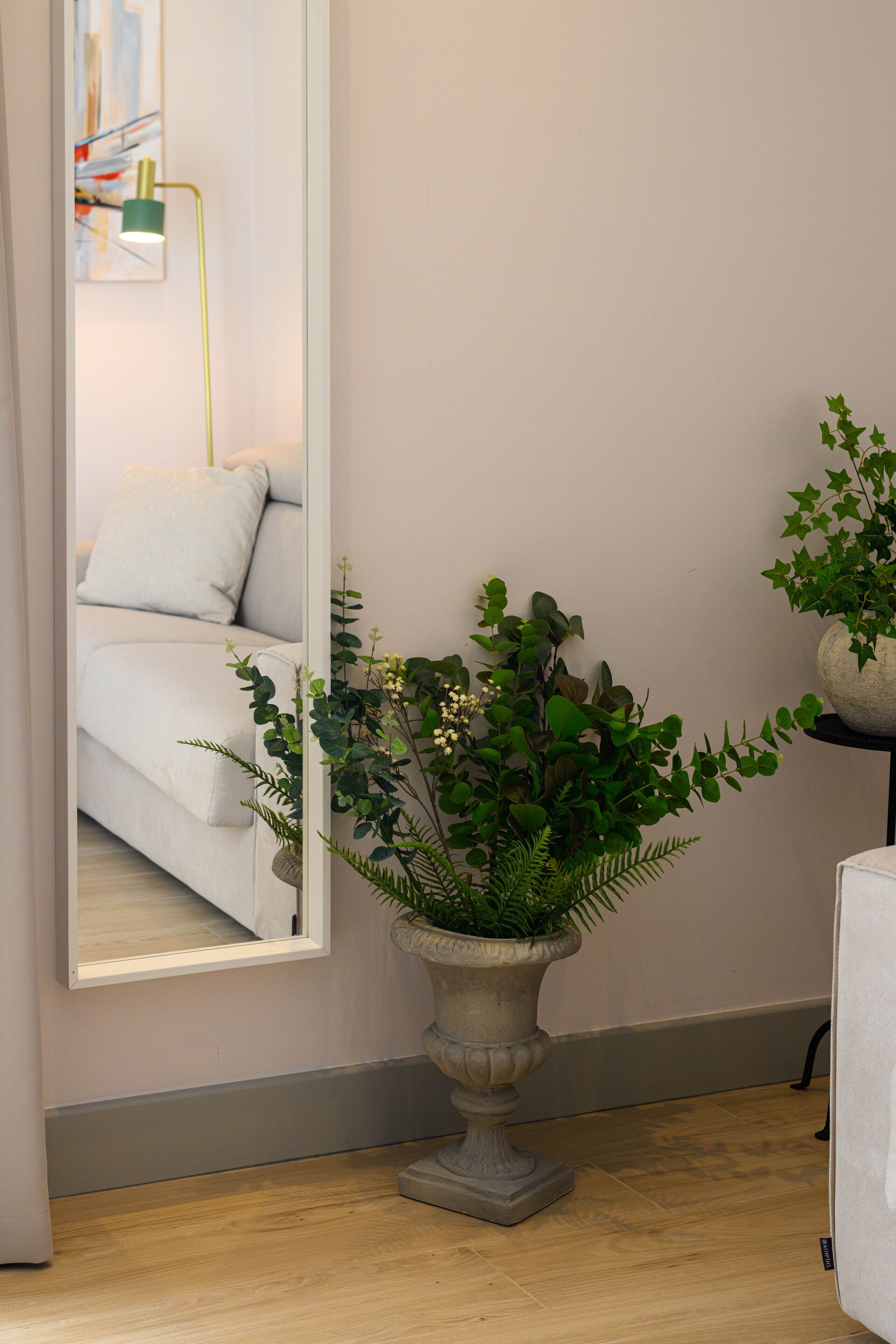 Une pièce dotée d’un grand miroir qui reflète un lit et une lampe de sol verte. On y trouve également une plante en pot, avec des feuilles vertes et des fleurs blanches, posée sur un plancher en bois.