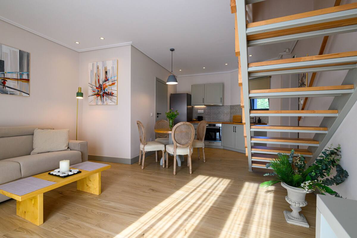Een moderne woonkamer met een bank, salontafel en een eethoek met stoelen. Een trap met houten leuningen leidt naar een hoger niveau.