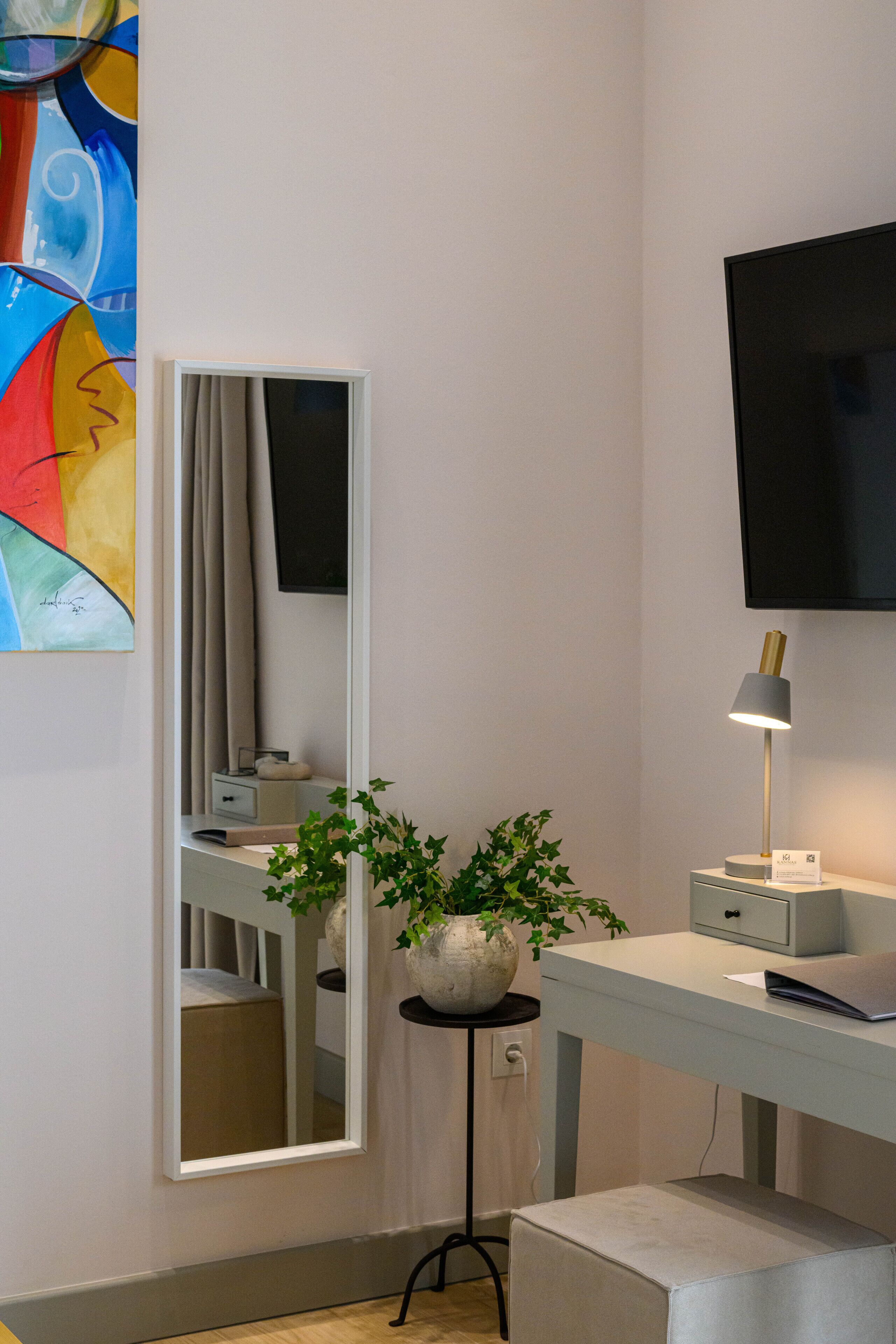 A modern room with a large mirror, a potted plant, a desk with a lamp, and a flat-screen TV mounted on the wall.