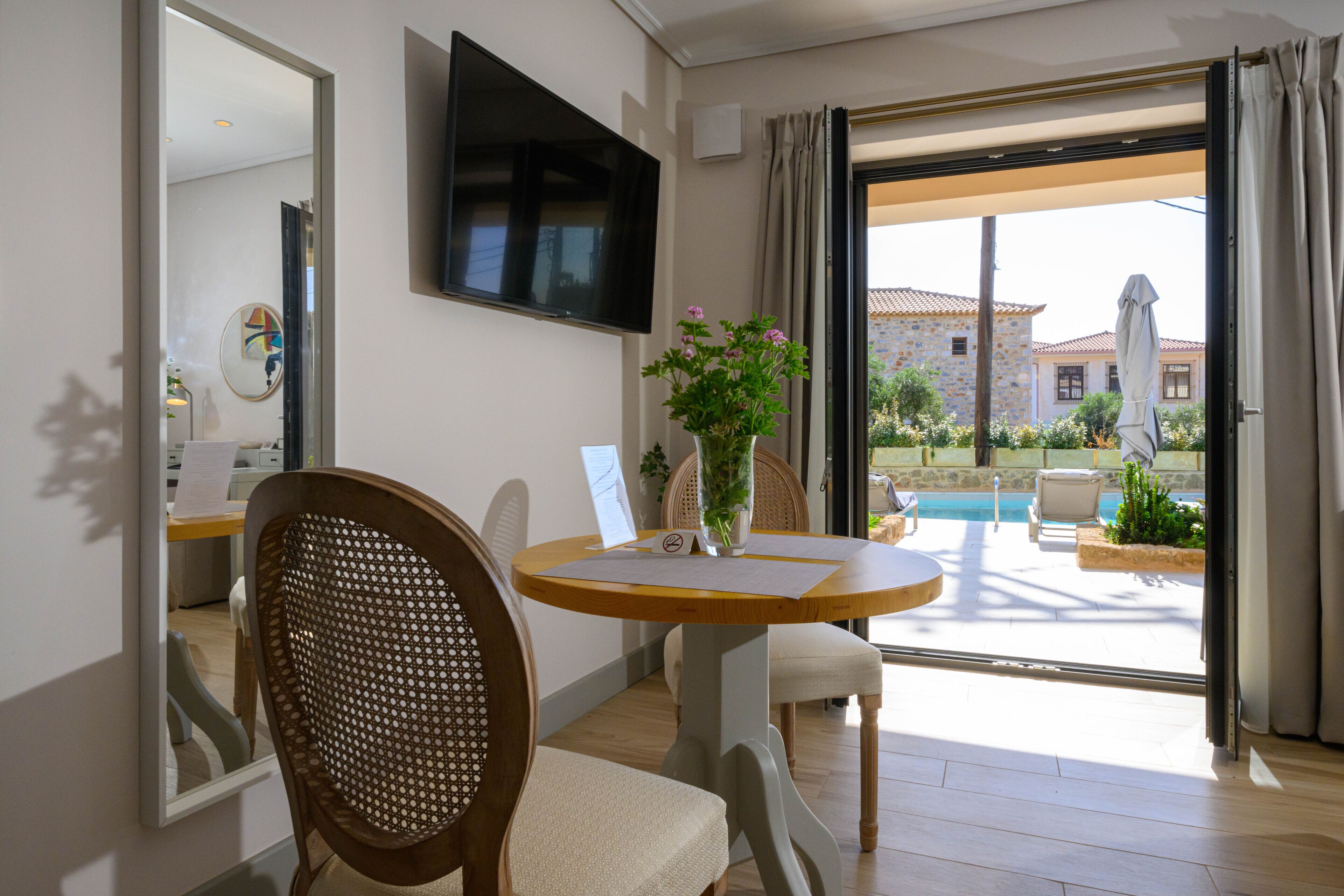 A hotel room with a dining table, chairs, a TV mounted on the wall, a view of the pool and outdoor seating area.