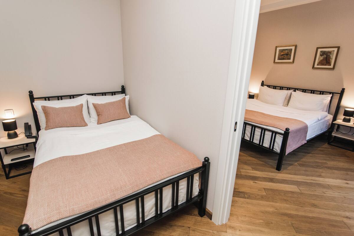 A bedroom with two beds, each with a nightstand and a lamp. The room has wooden flooring and white walls.
