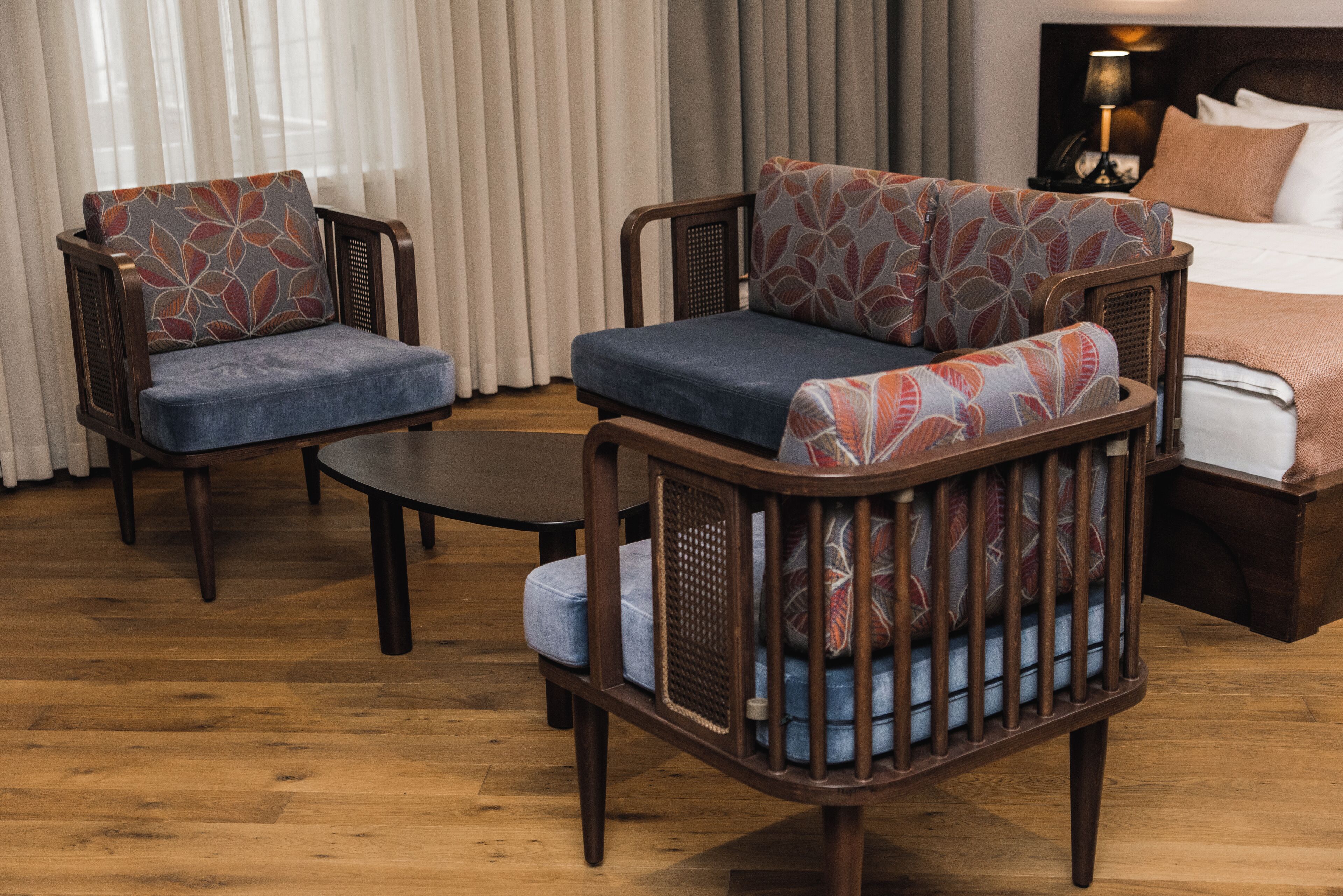 Habitación de hotel con suelo de madera, dos sillones con tapicería estampada, una mesita y una cama con cabecera.