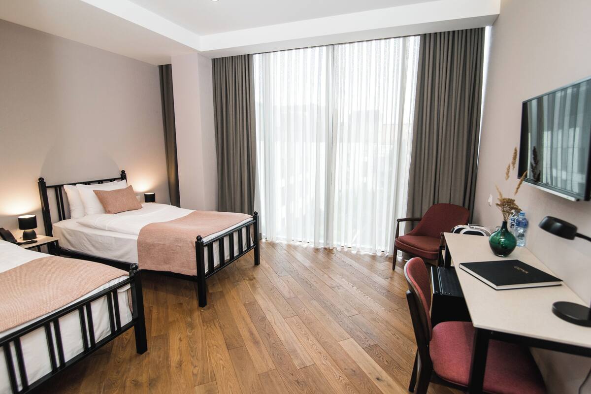 A hotel room with two beds, a desk, a chair, a TV, and a large window with curtains.
