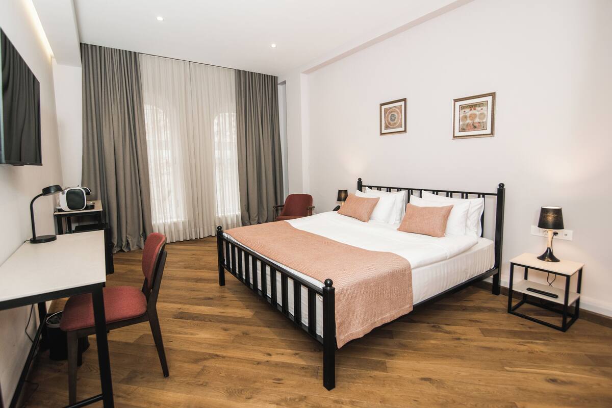 A bedroom with a bed, a desk, a chair, a bedside table, and a TV.