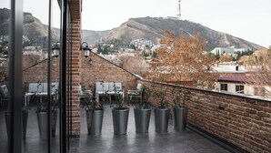 Varanda com plantas em vasos, parede de tijolos e vista para uma paisagem urbana com edifícios e colinas.