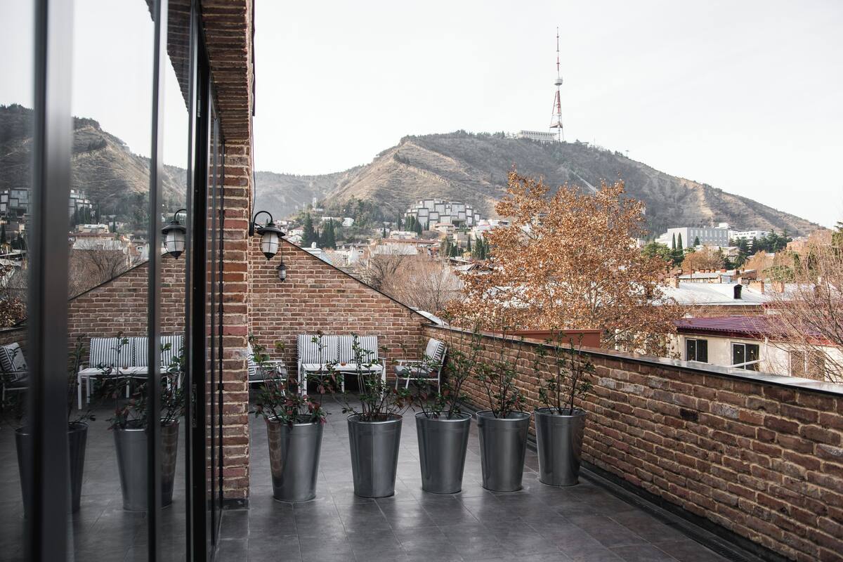A balcony with potted plants, a brick wall, and a view of a cityscape with buildings and hills.