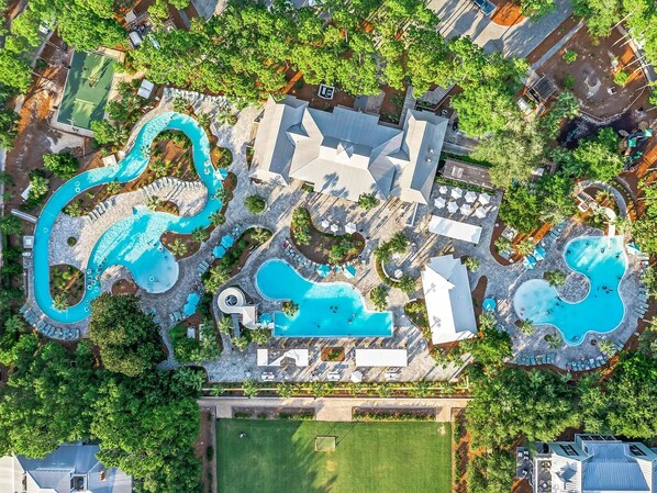 An aerial view of a resort with multiple swimming pools, a tennis court, and lush greenery.