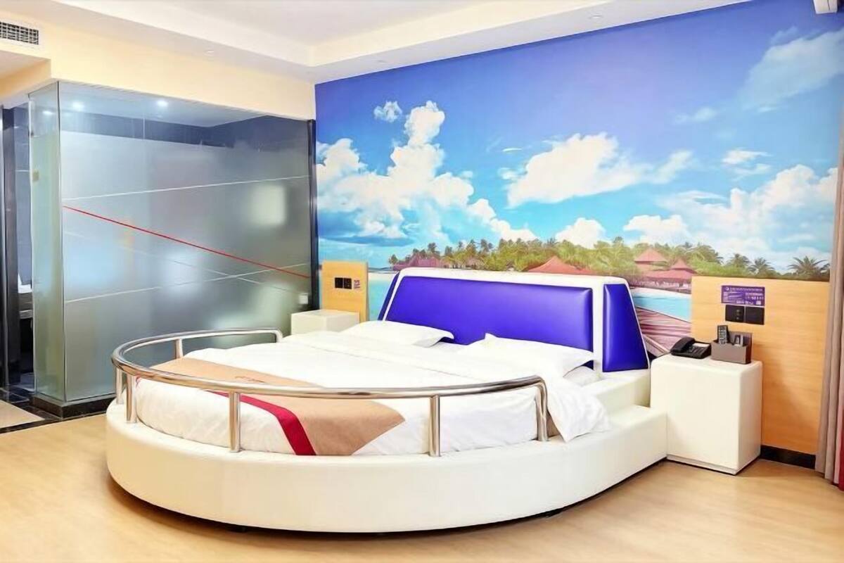 A circular bed with a white frame, a blue headboard, and a beige blanket. There is a reception desk with a wooden counter and a blue chair. A glass elevator is visible to the left.