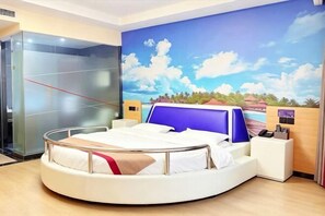 A circular bed with a white frame, a blue headboard, and a beige blanket. There is a reception desk with a wooden counter and a blue chair. A glass elevator is visible to the left. - Shankee Hotel (Shanghai Jinshan Wanda) (Shanghai)