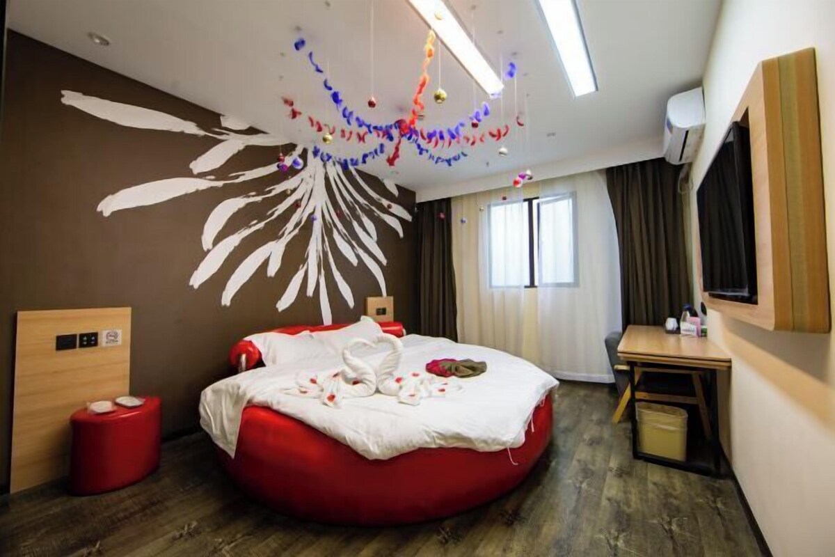 A hotel room with a red bean bag bed, a wooden headboard, a small desk, and a large window with curtains.