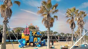 Un parc infantil a la platja amb palmeres, un camí de fusta i un paisatge urbà al fons.