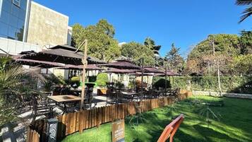 An outdoor seating area with tables and umbrellas, surrounded by a wooden fence and greenery.