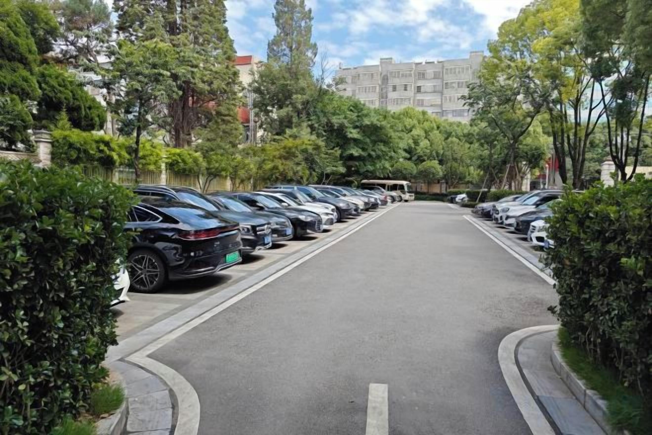A parking lot with multiple cars parked in rows, surrounded by greenery and buildings in the background.