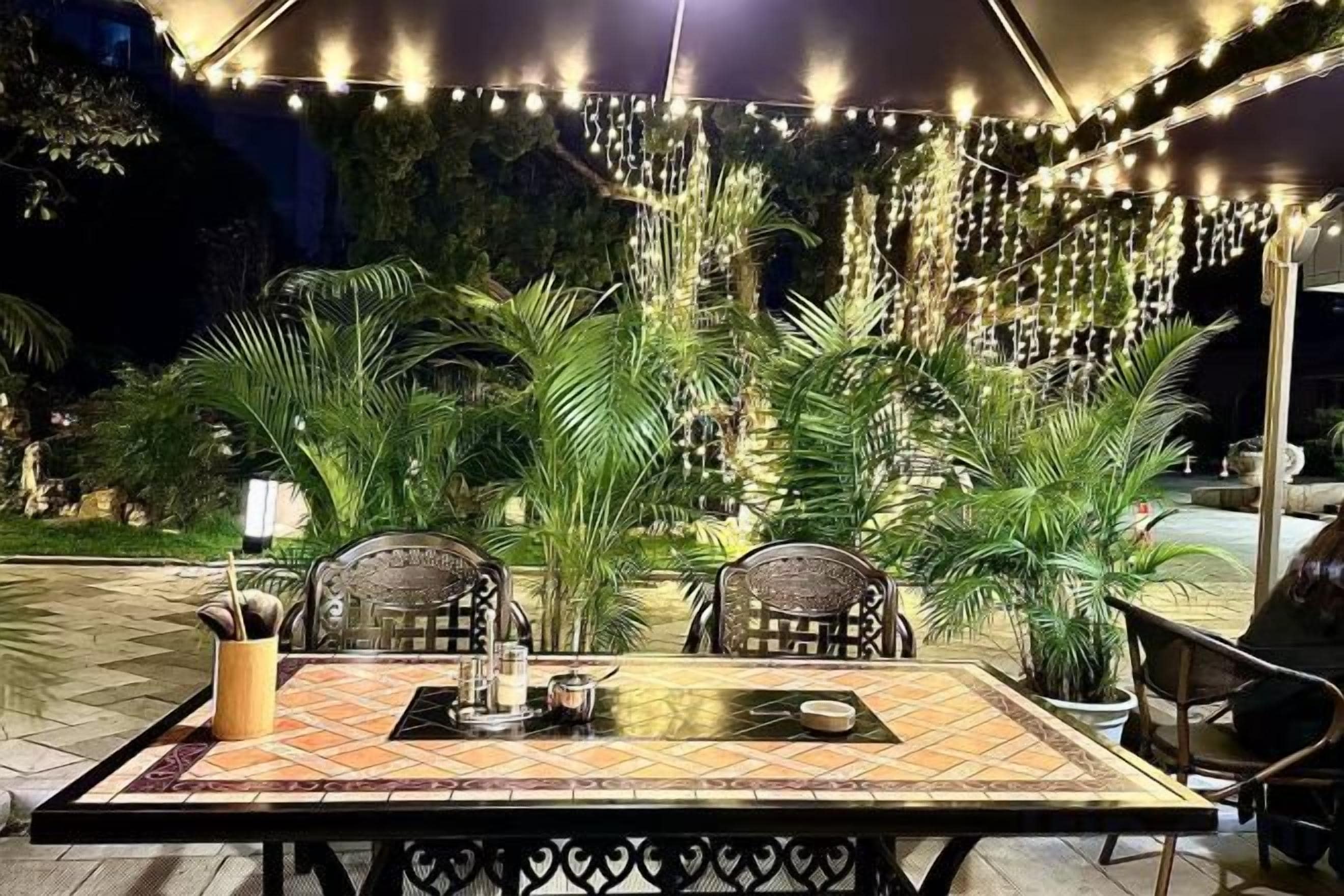 Un área de comedor al aire libre con una mesa y sillas, rodeada de plantas en macetas y luces colgantes.