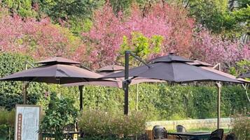 An outdoor seating area with tables and umbrellas, surrounded by greenery and flowering trees.