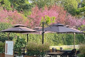 An outdoor seating area with tables and umbrellas, surrounded by greenery and flowering trees. - Lian Yun Hotel (Kunming)