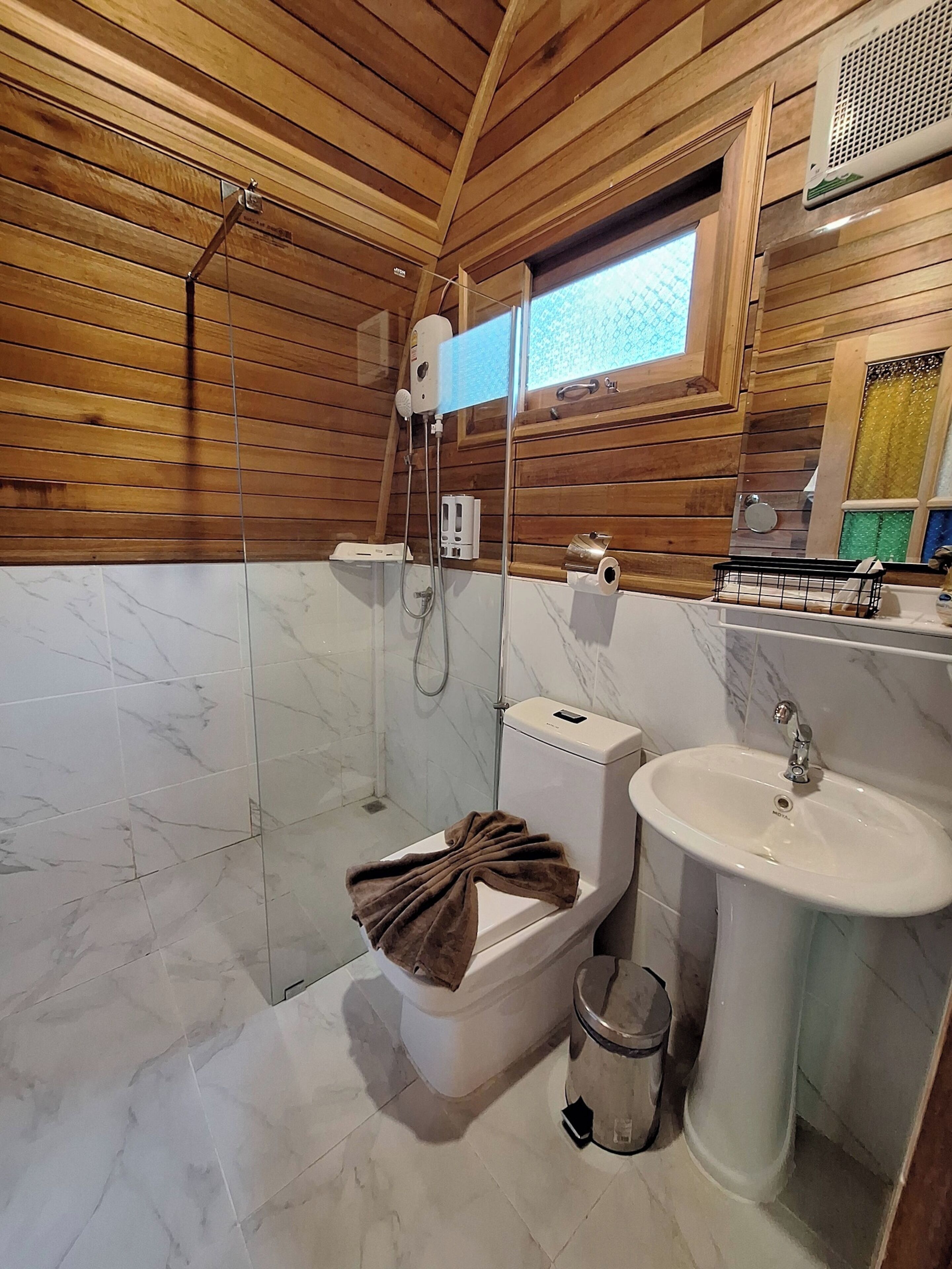 A bathroom with a wooden ceiling, a glass shower enclosure, a toilet, a sink, and a window with a view.