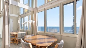 Ein geräumiges Zimmer mit einem großen Holztisch und Stühlen, großen Fenstern mit Meerblick und einer Stehlampe.