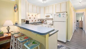 Una cocina con armarios blancos, encimera de granito, refrigerador, microondas y un lavamanos. Hay taburetes con diseños coloridos y una lámpara sobre la encimera.