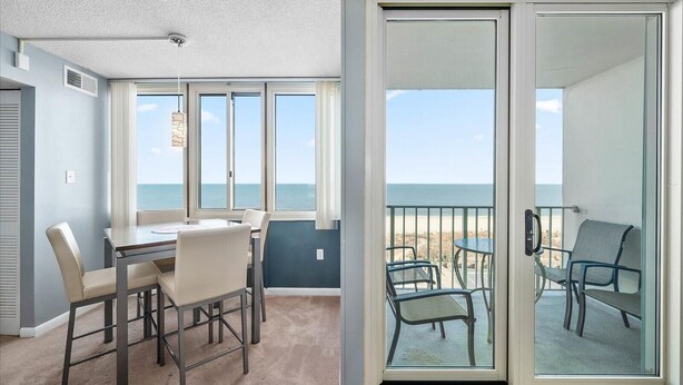 A room with a dining table and chairs, a sliding glass door leading to a balcony with chairs, and a view of the ocean. - Quay - Direct Ocean Front 3 Bedroom Condo with Indoor & Outdoor Pools! (Ocean City)