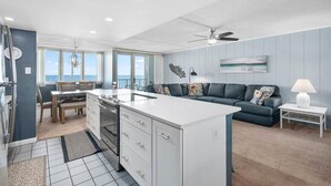 A modern kitchen with a central island, a dining area with a view, and a living space with a sofa and a lamp. - Quay - Direct Ocean Front 3 Bedroom Condo with Indoor & Outdoor Pools! (Ocean City)
